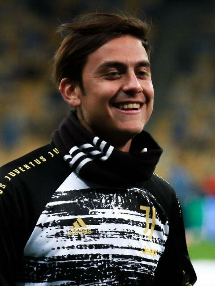 Paulo Dybala smiling during a warm-up session wearing Juventus training gear and a black scarf outdoors.