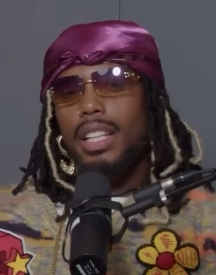 B.o.B speaking into a microphone wearing sunglasses and a purple headscarf, sharing bio and career highlights.