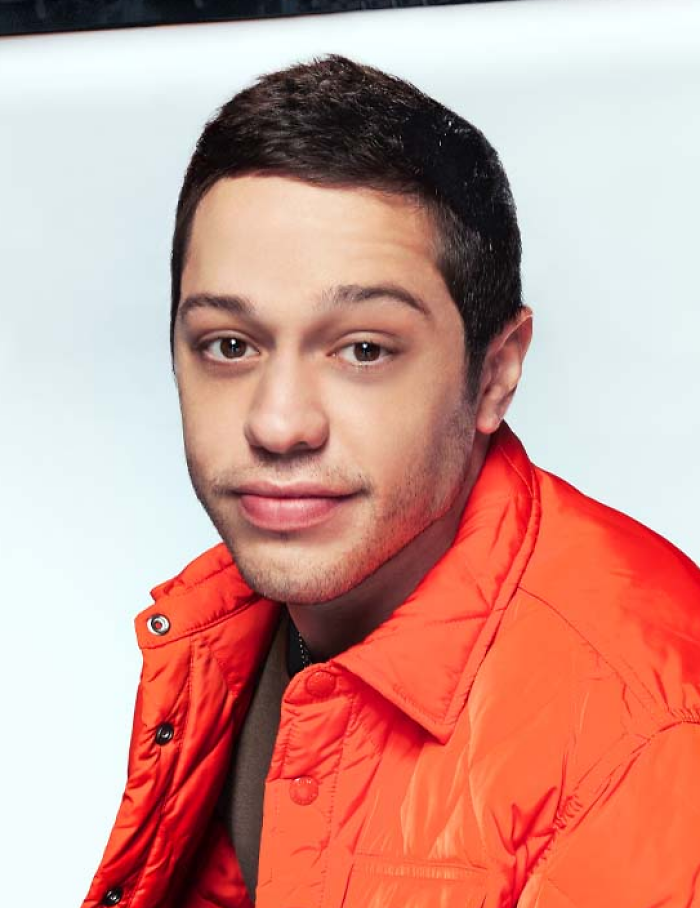 Pete Davidson wearing an orange jacket, looking at the camera with a slight smile in a studio setting.