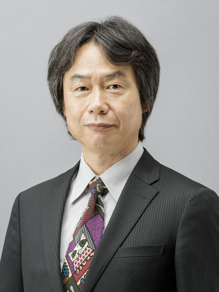 Shigeru Miyamoto wearing a dark suit and colorful tie, posing against a plain gray background in a professional portrait.