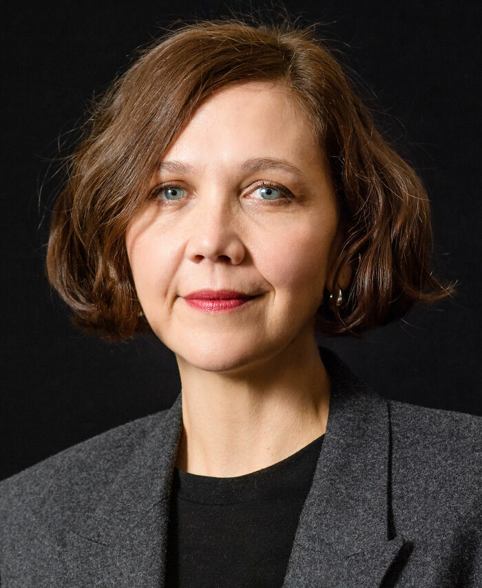 Maggie Gyllenhaal with short brown hair and red lipstick wearing a grey blazer against a black background