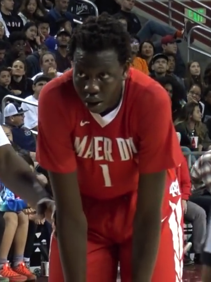 Bol Bol in a red basketball uniform during a game with spectators in the background at an indoor court.