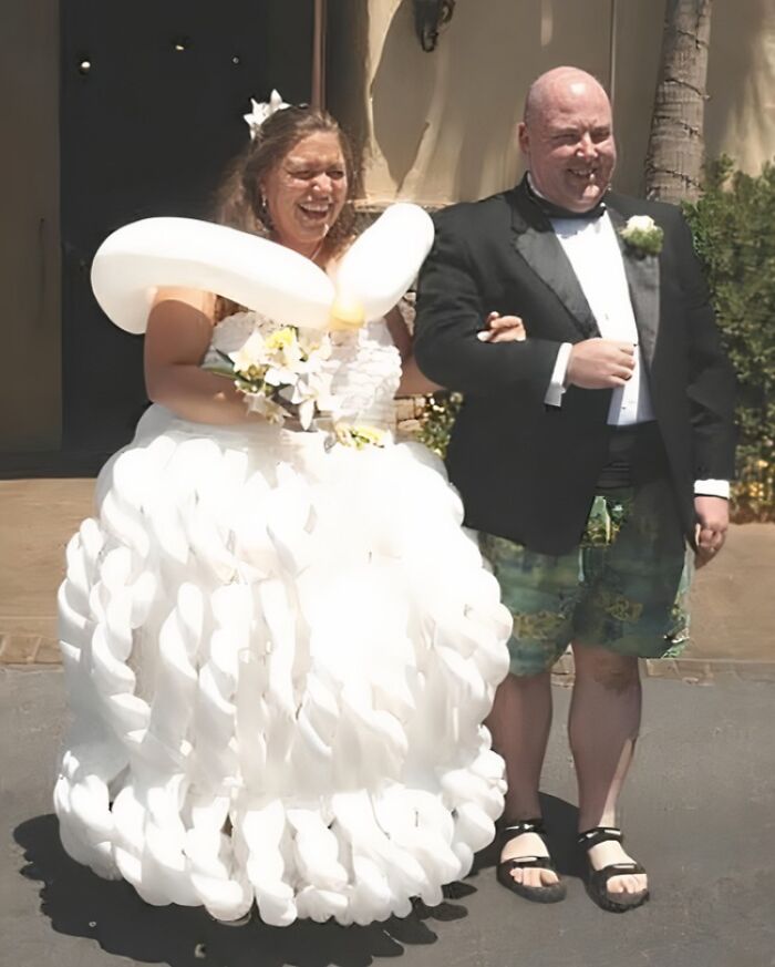Bride wearing a wedding dress made of white balloons standing next to a groom in a tuxedo jacket and shorts.