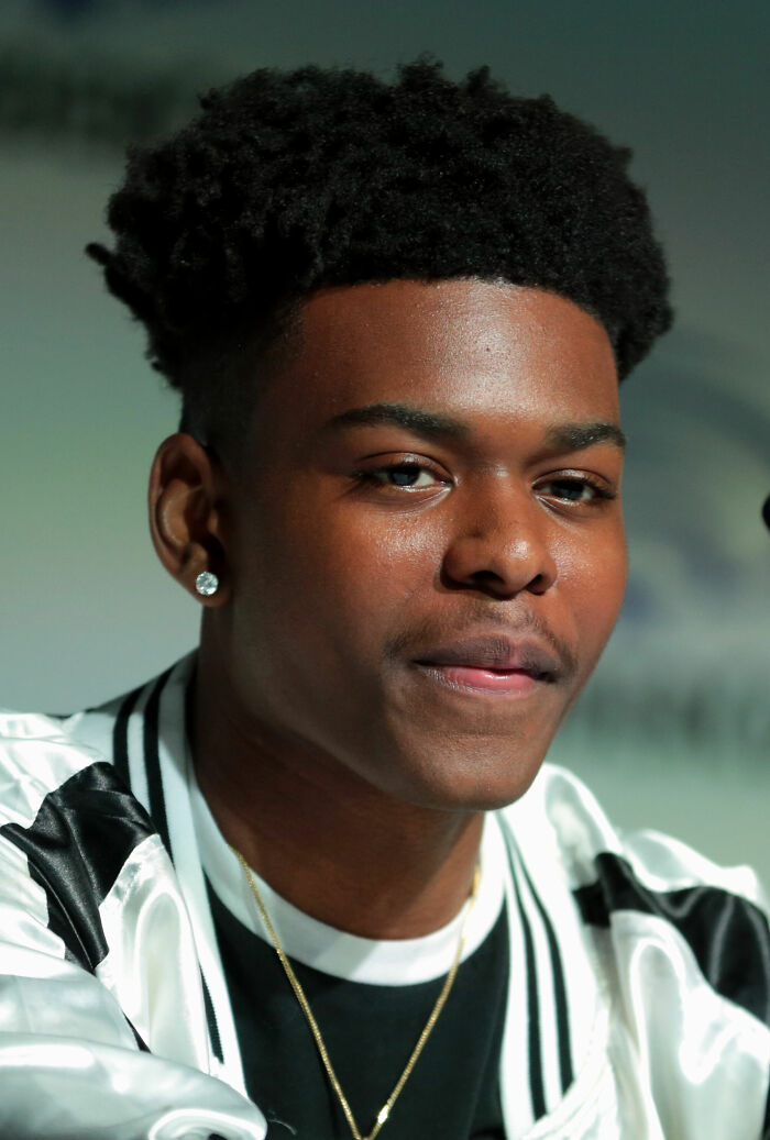 Aubrey Joseph close-up portrait wearing a white jacket and gold chain, showcasing his hairstyle and earring indoors.