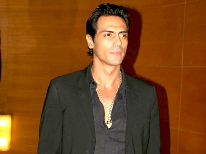 Arjun Rampal wearing a black suit and chain necklace, posing confidently against a wooden background.