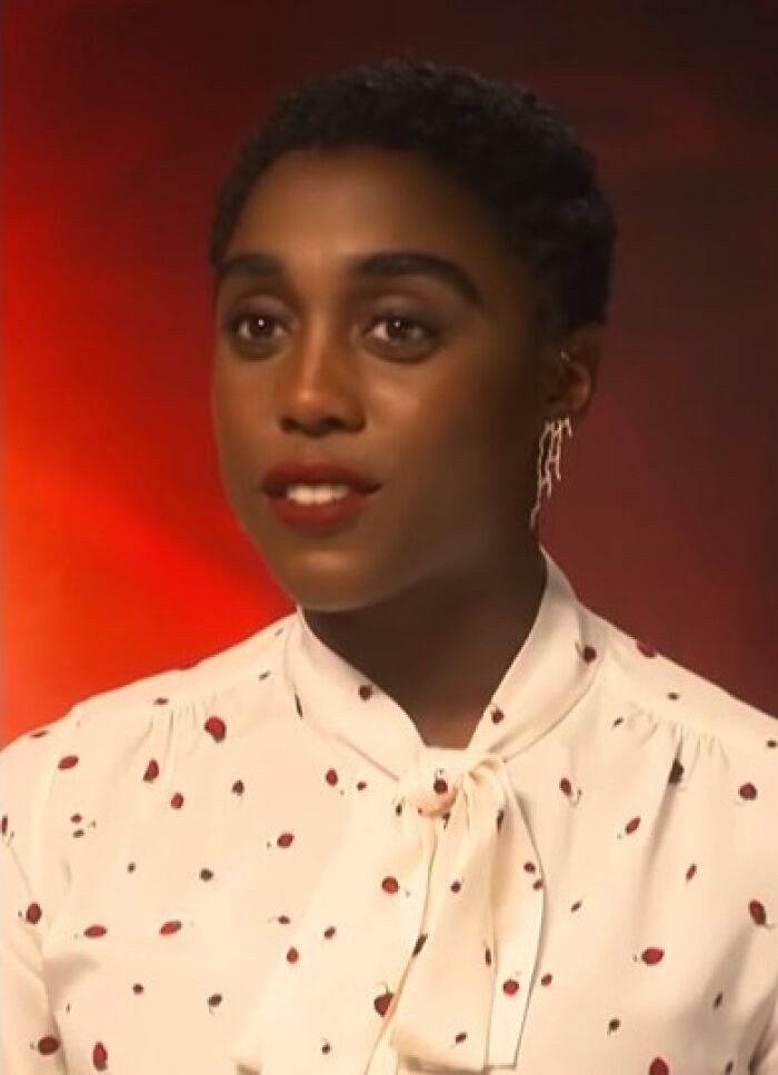 Lashana L***h wearing a white blouse with red spots and unique earrings, speaking against an orange background.