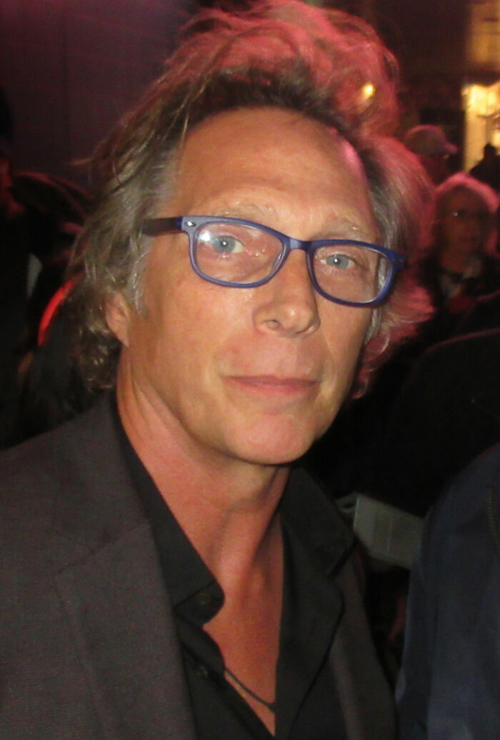 William Fichtner wearing glasses and a dark jacket at a public event with blurred background crowd.
