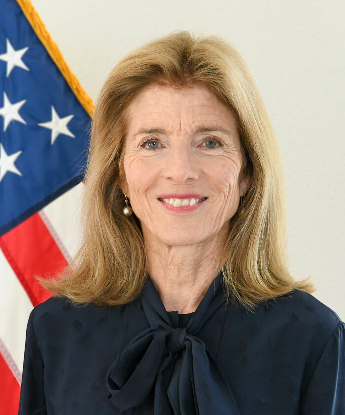 Caroline Kennedy smiling in front of an American flag, wearing a navy blouse with a bow, highlighting her bio and career.
