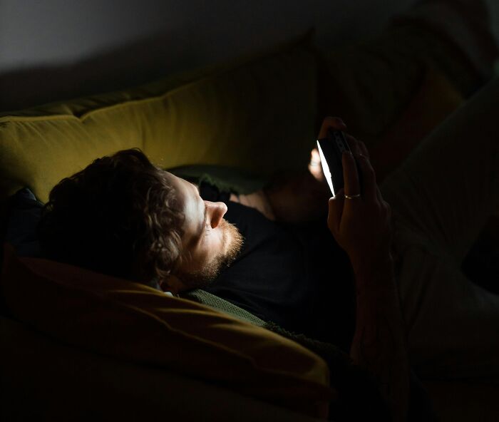 Man lying on couch scrolling phone in dark room, relatable behavior illustrating ridiculous but relatable things.