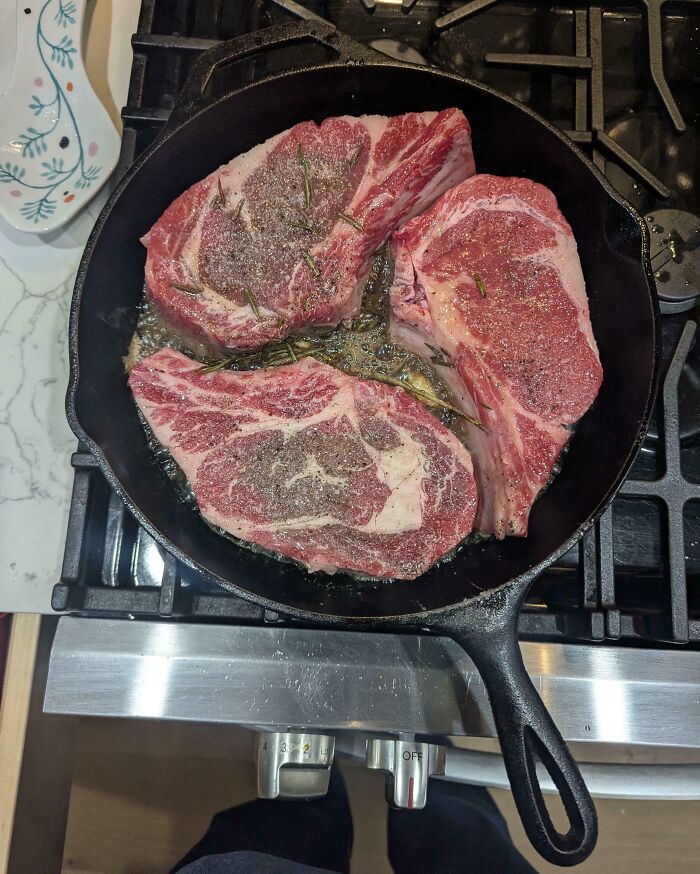 Three perfectly aligned seasoned steaks cooking in a cast iron skillet on a stovetop burner.