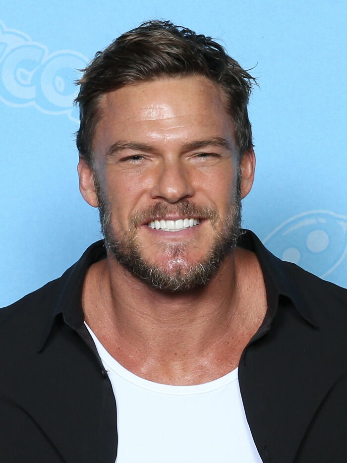 Alan Ritchson smiling at an event, wearing a black shirt over a white tee against a blue background.