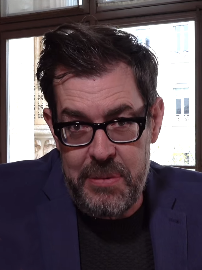 Richard Osman wearing glasses and a blazer, sitting indoors near a window, highlighting his bio and career.