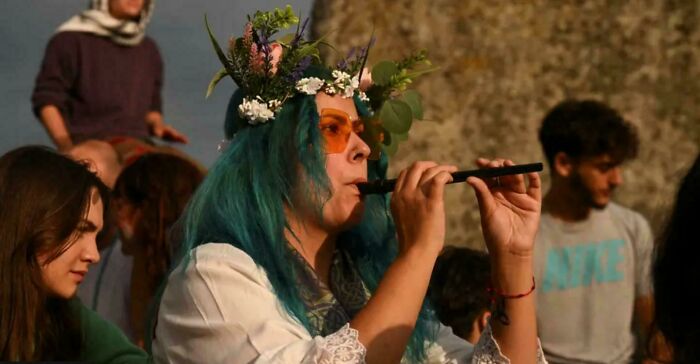 Woman with blue hair and floral crown playing a flute, showcasing beauty and diversity in an outdoor gathering portrait.