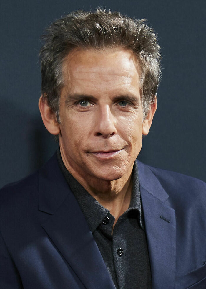 Ben Stiller wearing a dark shirt and blazer, posing with a slight smile against a dark background.