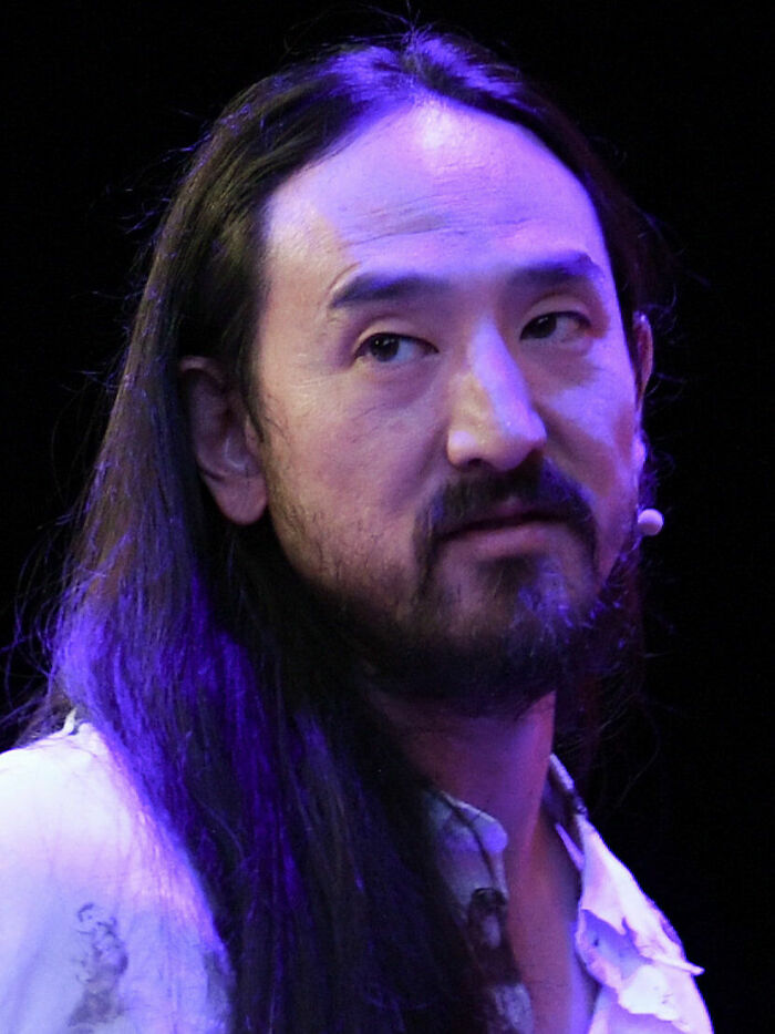 Steve Aoki speaking on stage with long hair and beard, highlighted by purple lighting in a close-up portrait.