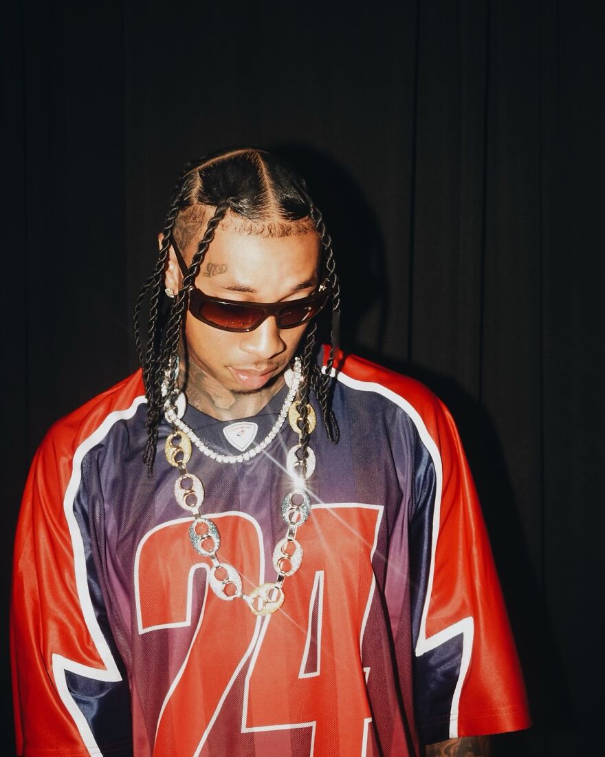 Tyga with braided hair and sunglasses wearing a red and blue jersey, featured on members-only fan pages.