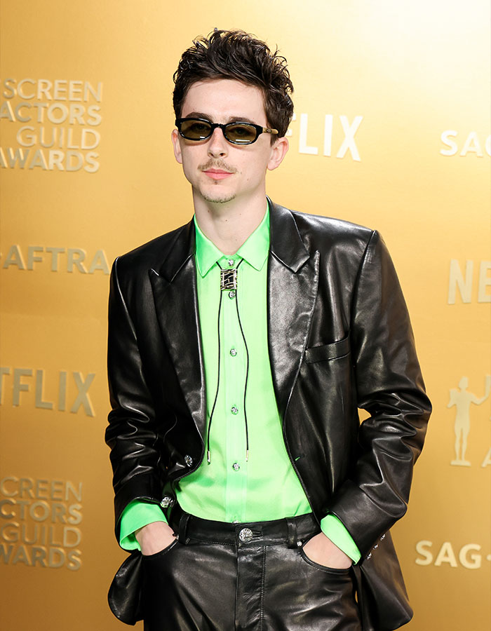 Timothée Chalamet wearing a black leather suit with a neon green shirt and sunglasses at an awards event. Timothée Chalamet wearing a black leather suit with a neon green shirt and sunglasses at an awards event.