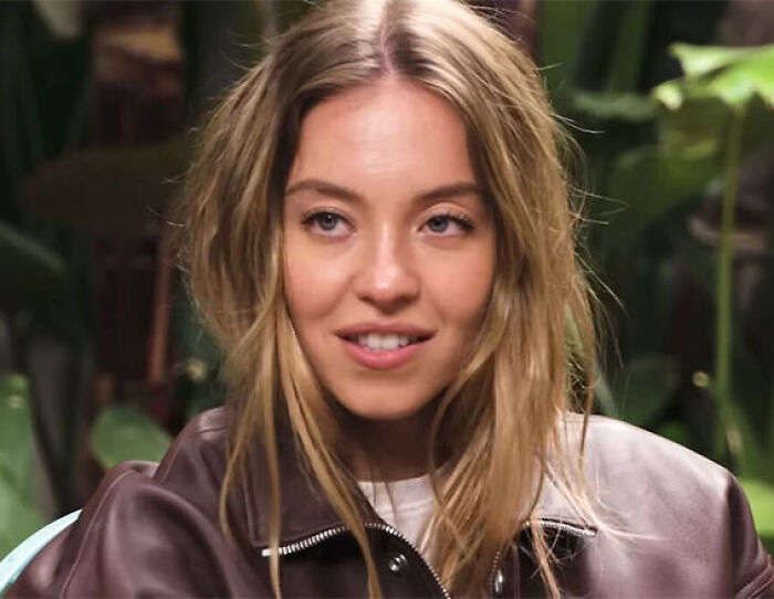 Young woman with blonde hair wearing a brown leather jacket, smiling subtly during a casual indoor interview setting.