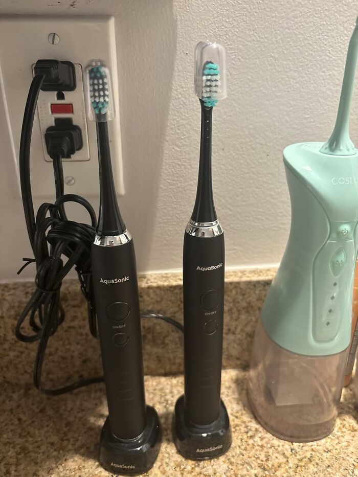 Two black AquaSonic electric toothbrushes on charging stands beside a water flosser on a bathroom countertop, holiday shopping gift idea.