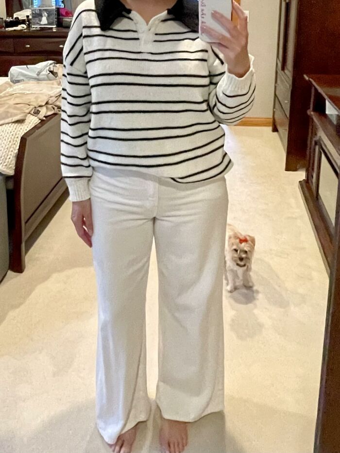 Person wearing white striped top and white pants taking mirror selfie with small dog in background, Alix Earle Amazon store deals.