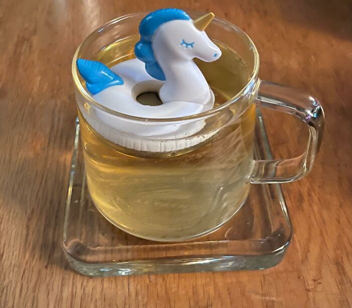 Unicorn-shaped novelty tea infuser floating in a glass cup filled with tea on a wooden surface.