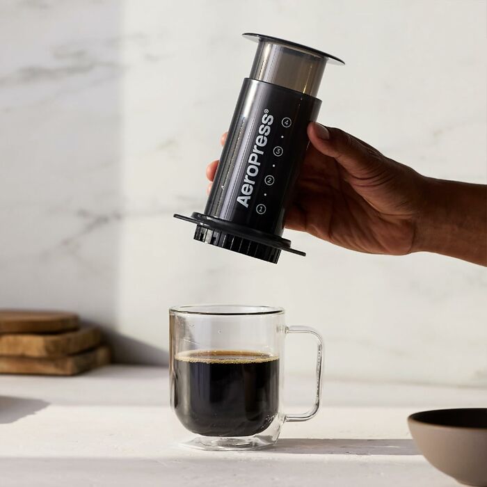 Hand holding an Aeropress coffee maker over a glass cup of black coffee, a foolproof find for gift shoppers.
