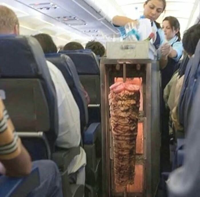 Airplane cabin with flight attendants serving while a large vertical meat rotisserie stands in the aisle, a go hard pic liked most.