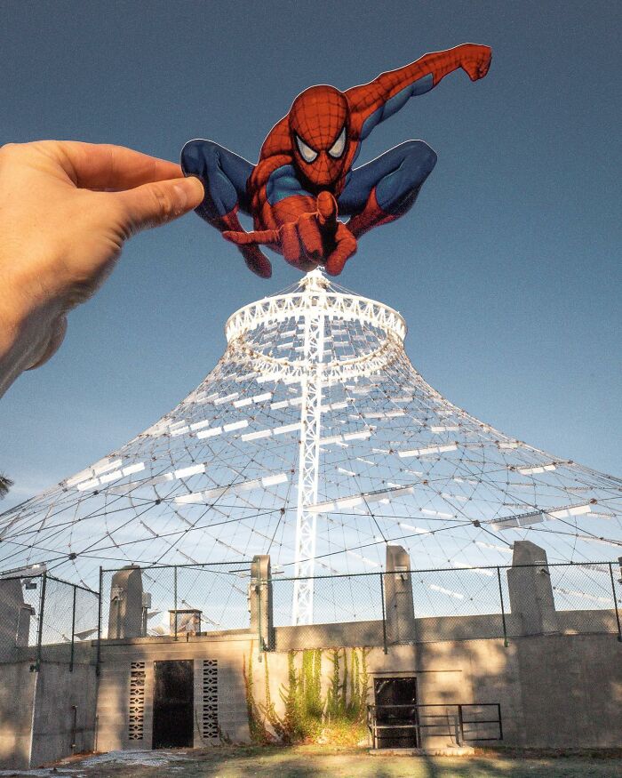 Hand holding paper cutout of Spider-Man appearing to interact with a white tower structure in daylight, paper cutouts. Hand holding paper cutout of Spider-Man appearing to interact with a white tower structure in daylight, paper cutouts.
