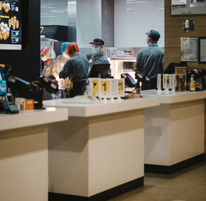Fast food workers wearing masks and uniforms preparing orders behind counter in busy restaurant kitchen.