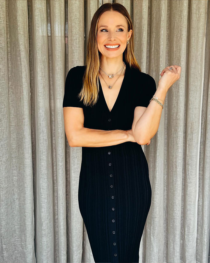 Kristen Bell smiling in a black dress with layered necklaces and bracelets, standing against a beige curtain backdrop.