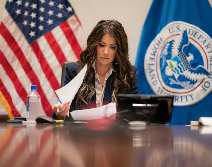 Kristi Noem working at a desk with American flag and Homeland Security emblem in the background, showing transformation.