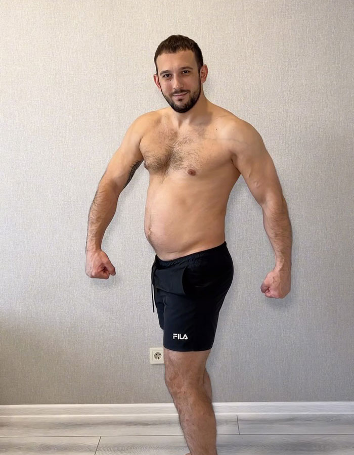 Shirtless fitness coach posing indoors wearing black shorts, showing muscular arms and slight belly against plain wall background. Shirtless fitness coach posing indoors wearing black shorts, showing muscular arms and slight belly against plain wall background.