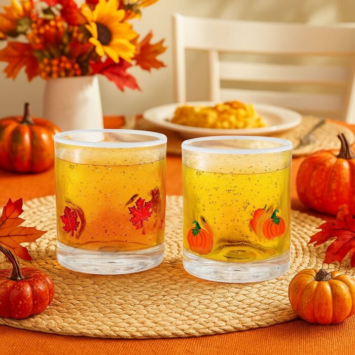 Two festive glasses with fall leaf and pumpkin designs on a woven placemat surrounded by autumn decor.