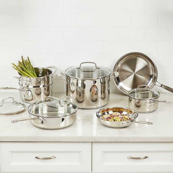 Stainless steel kitchen cookware set displayed on countertop including pots, pans, lids, and steamer with asparagus.