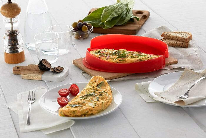 Silicone omelette cooker with cooked omelette, cherry tomatoes, fresh spinach, and kitchen finds on white table.