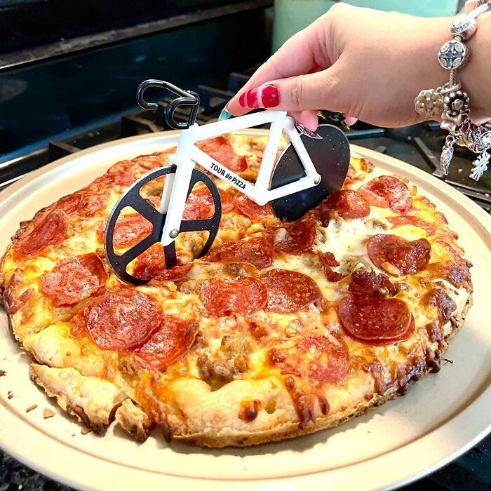 Hand holding a bicycle-shaped pizza cutter slicing a pepperoni pizza, showcasing a unique novelty finds kitchen tool.