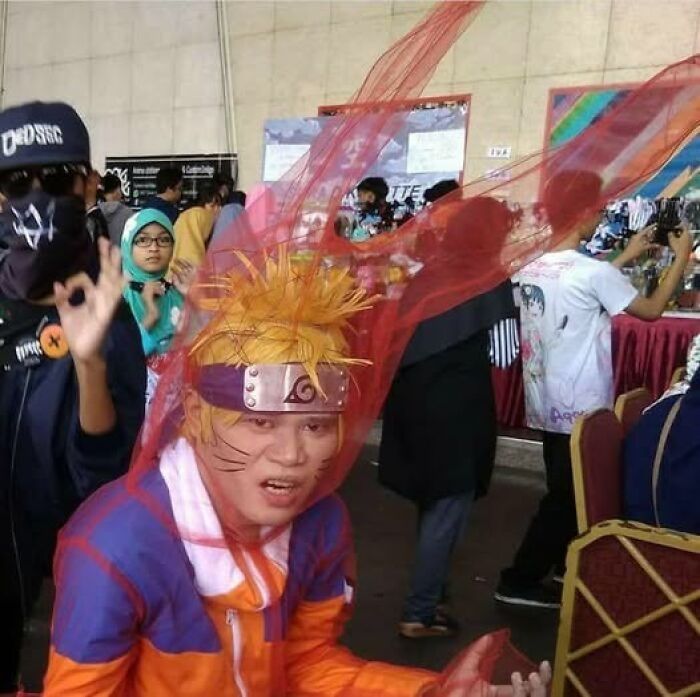 Cosplayer dressed as Naruto in an orange costume with a red scarf at a crowded event for pics that go hard.