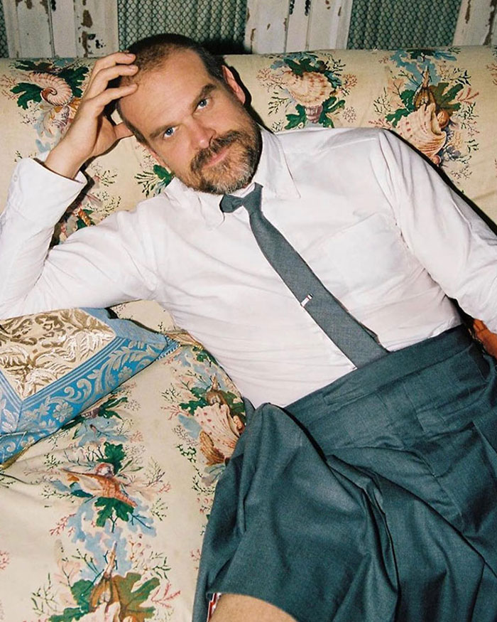 Man with a beard reclining on floral couch in white shirt and gray tie, contrasting Lily Allen’s CFDA fashion look debate online.