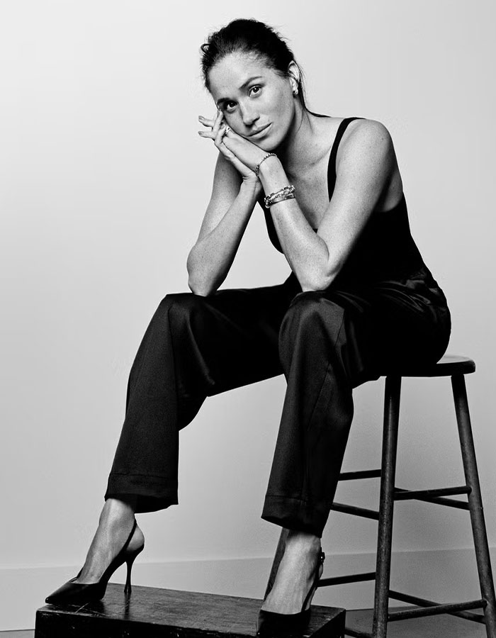 Meghan Markle sitting on a stool in black outfit and heels, posing thoughtfully in a black and white editorial photo. Meghan Markle sitting on a stool in black outfit and heels, posing thoughtfully in a black and white editorial photo.