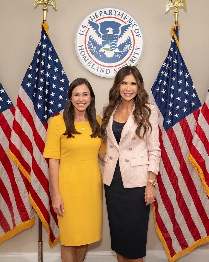 Two women posing in front of U.S. Department of Homeland Security sign with American flags, showcasing Kristi Noem transformation.