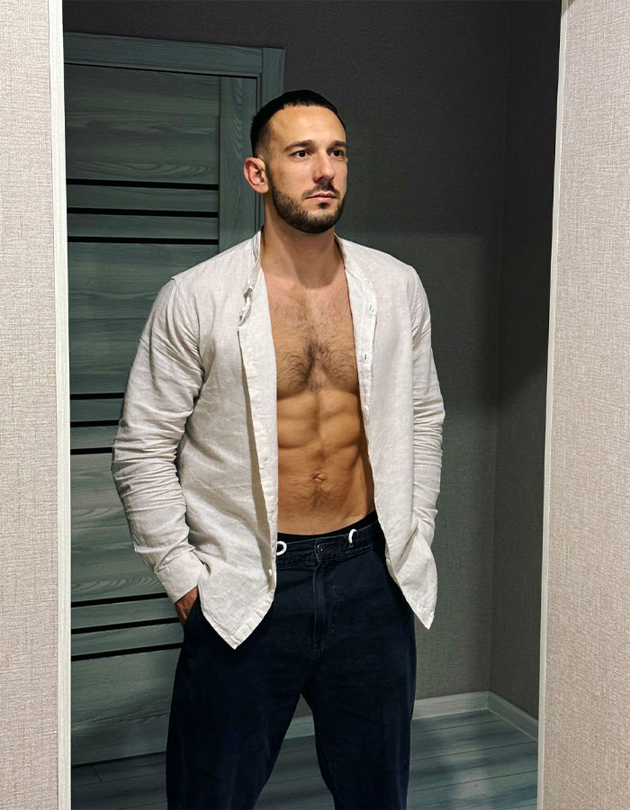 Fitness coach with visible abs wearing an open white shirt and dark pants standing indoors against a gray wall. Fitness coach with visible abs wearing an open white shirt and dark pants standing indoors against a gray wall.