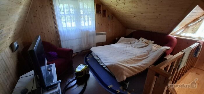 Cozy attic bedroom with wooden walls and ceiling, mismatched furniture, and a vacuum cleaner on the floor in a wild real estate listing.