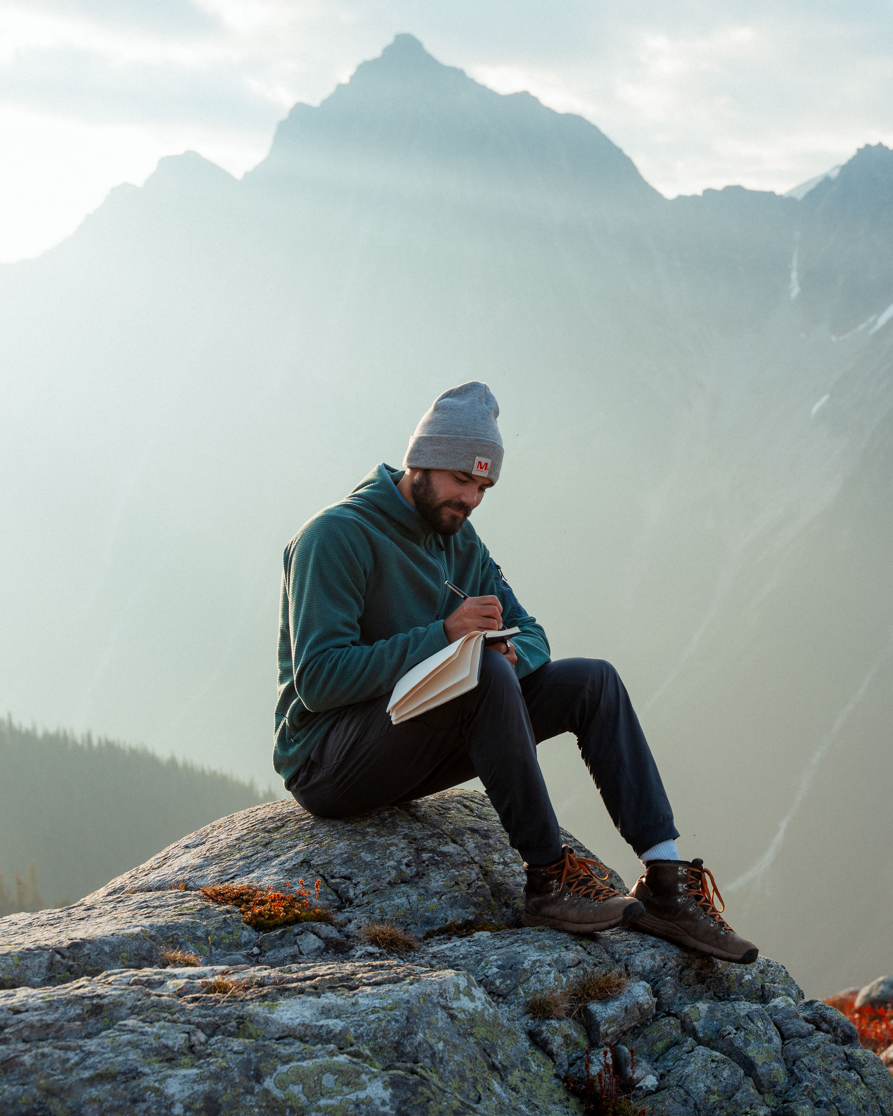 Man writing in journal on mountain rock, reflecting on purpose while surrounded by misty peaks during outdoor adventure Man writing in journal on mountain rock, reflecting on purpose while surrounded by misty peaks during outdoor adventure