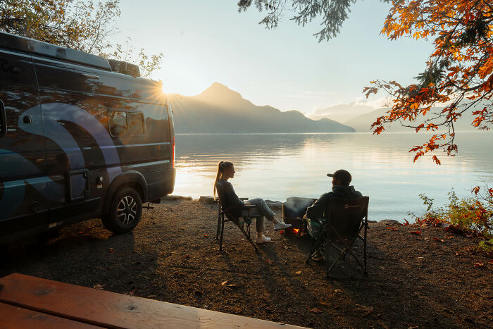Two people sitting by a campfire near a van, reflecting on their journey and purpose beside a calm lake at sunset. Two people sitting by a campfire near a van, reflecting on their journey and purpose beside a calm lake at sunset.