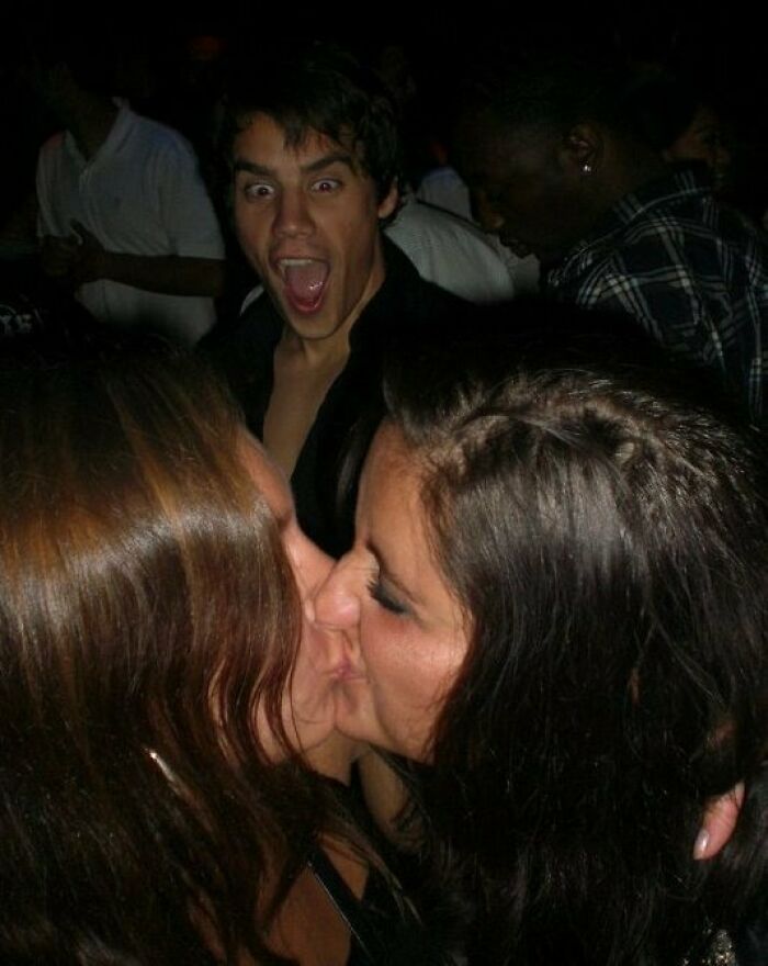 Young man photobombing with a surprised face behind two women kissing at a lively party setting.