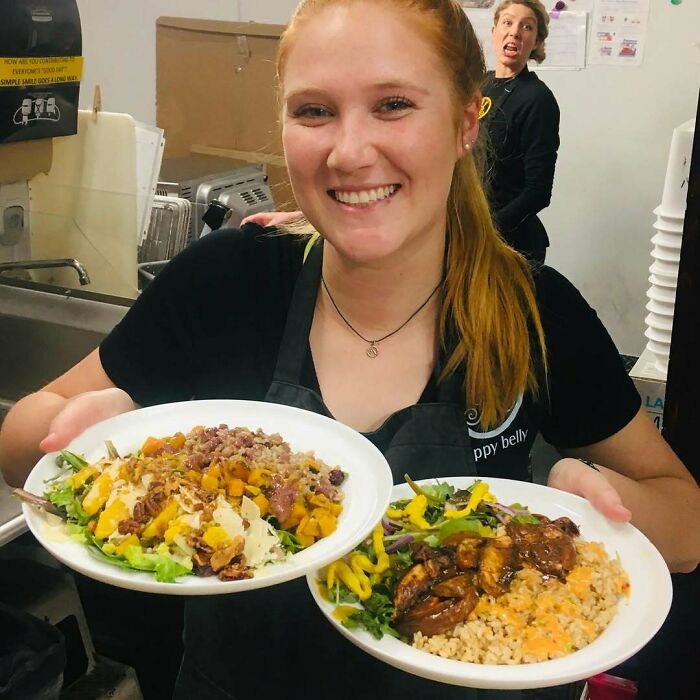 Young woman smiling while holding two plates of food with a funny photobomb in the background, epic photobombs theme.