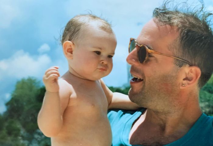 Man wearing sunglasses holding a baby outdoors with blue sky, related to Bruce Willis dementia update keywords. Man wearing sunglasses holding a baby outdoors with blue sky, related to Bruce Willis dementia update keywords.
