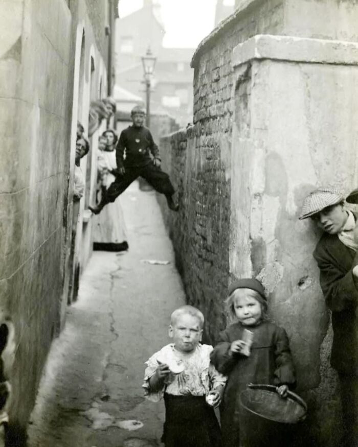 Children and adults in a narrow Victorian era alley, with a boy suspended between walls, showcasing weird photos from the Victorian era.