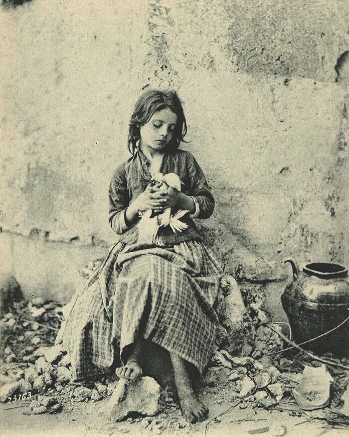 Victorian era photo of a barefoot girl sitting on rubble holding a bird, showing the different time and lifestyle of that era.