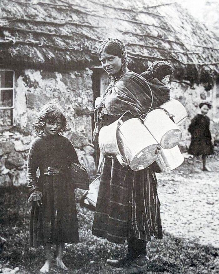 Victorian era woman carrying milk cans with children outdoors near a stone cottage, a weird photo showcasing a different time.